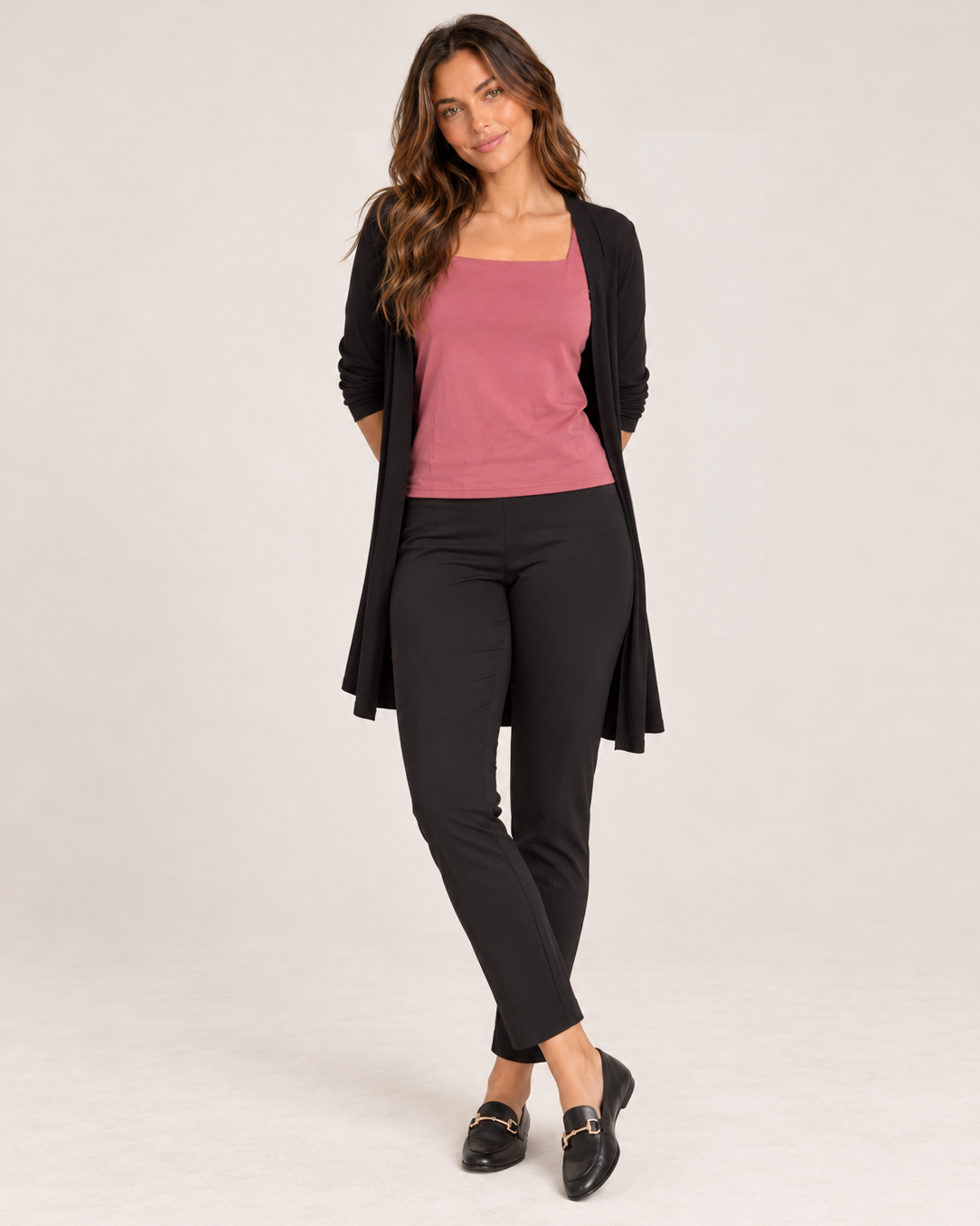 Full-body front view of a woman wearing a muted pink square-neck short-sleeve fitted top with black pants and a black open-front cardigan on a beige background.