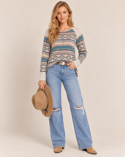 Full-body front view of a woman wearing a multicolor Southwestern geometric-pattern knit sweater with light-wash jeans and a tan wide-brim hat on a beige background.