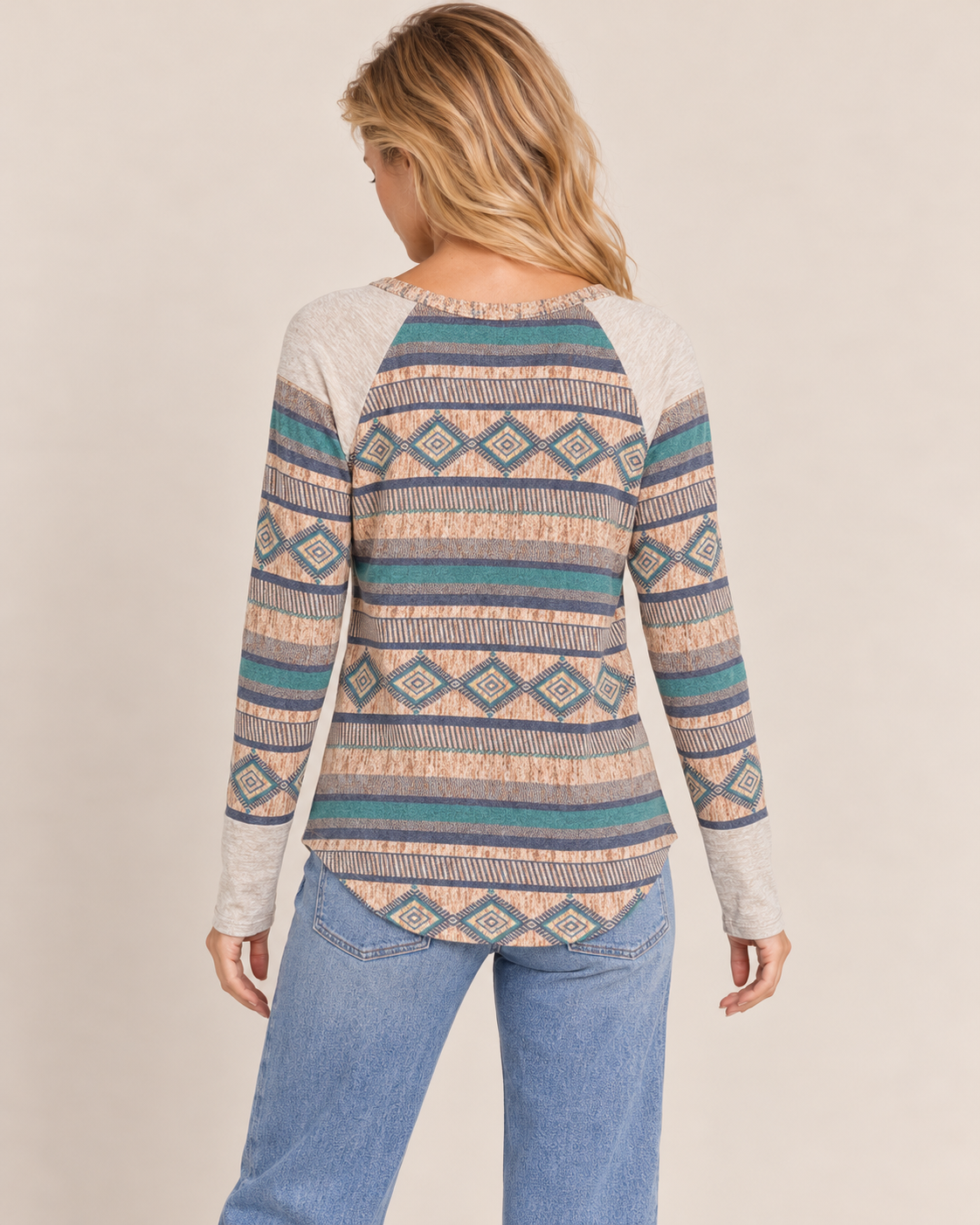 Back view of a woman wearing a multicolor Southwestern geometric-pattern knit sweater featuring horizontal bands of diamond and zigzag motifs in teal, rust, cream, and gray on a beige background.