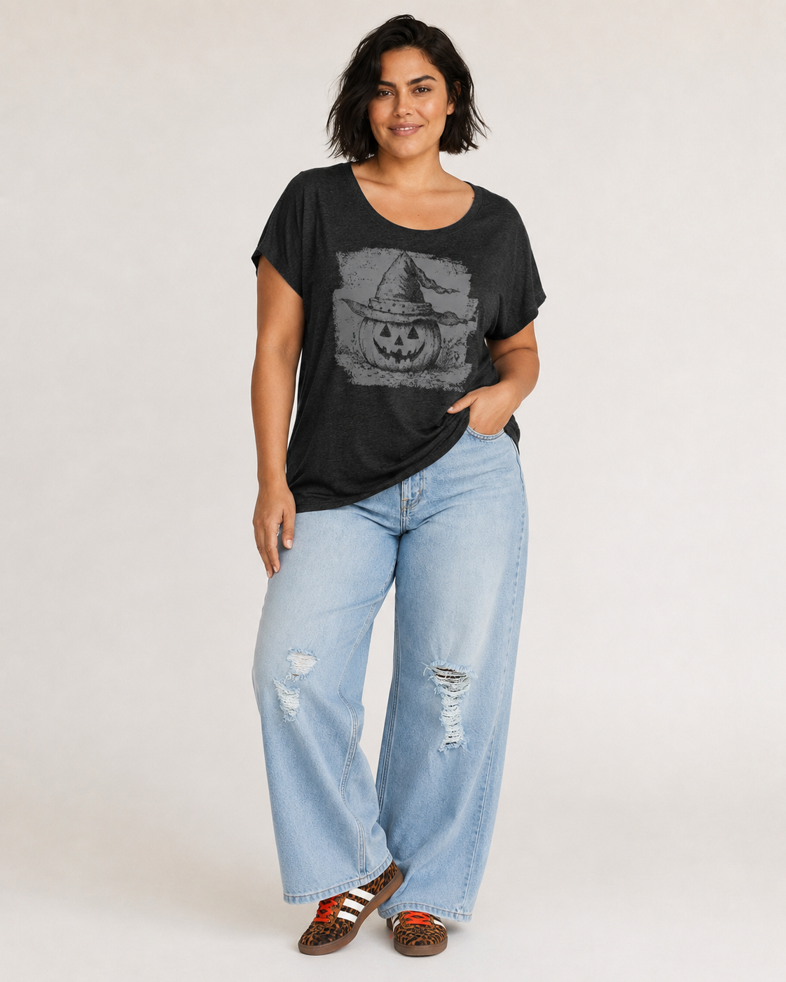 Full-body view of a woman wearing a black pumpkin graphic tee with light blue jeans and black shoes on a beige background