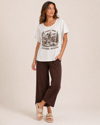 Full-body view of a woman wearing a navy graphic tee featuring a "Peace Comes in Waves" design, light wash denim shorts, and white sneakers on a beige background