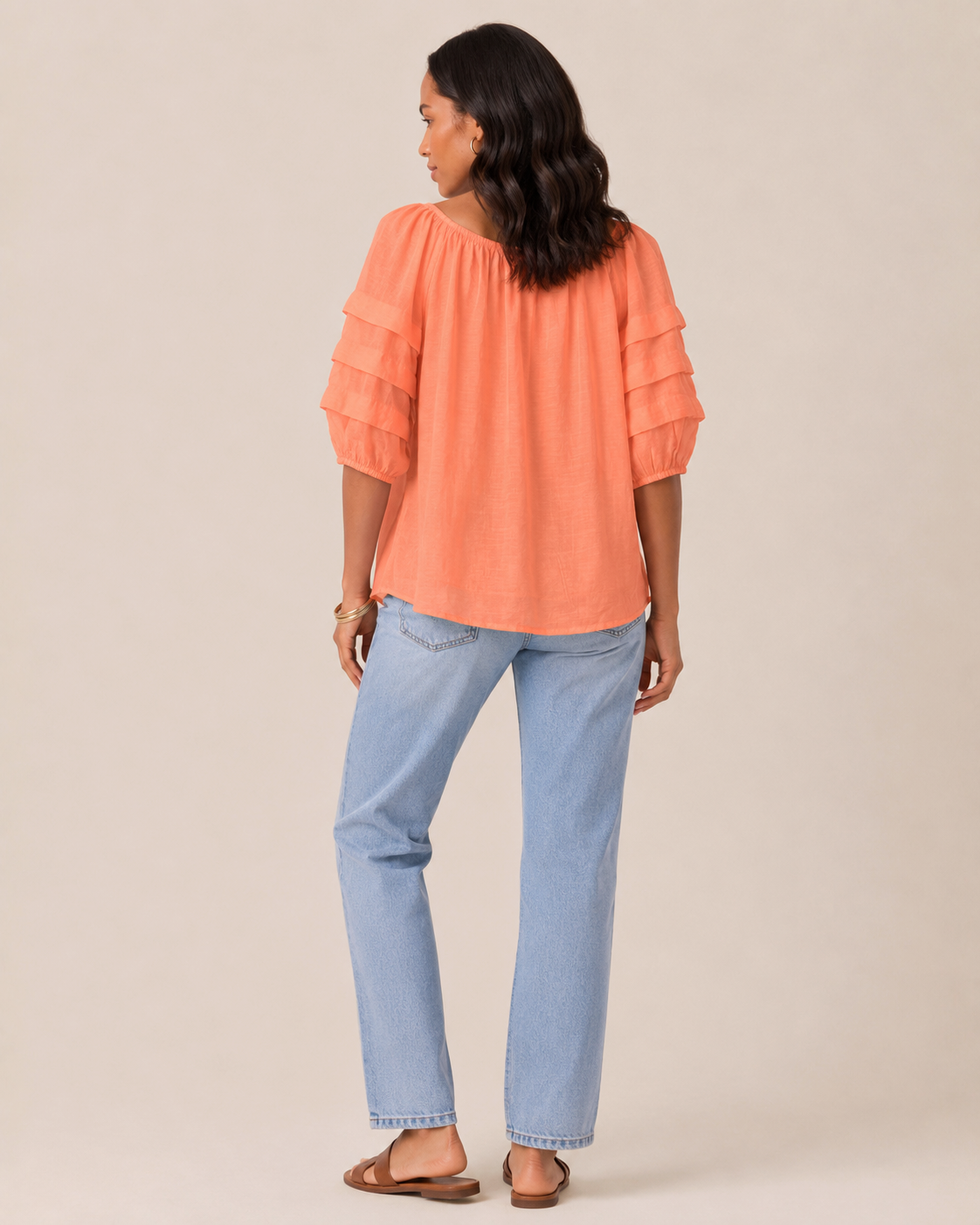 Back view of a woman wearing a cantaloupe tier sleeve top and light wash denim jeans on a beige background