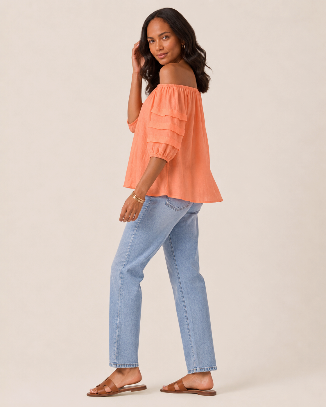 Side view of a woman wearing a cantaloupe tier sleeve top and light wash denim jeans on a beige background