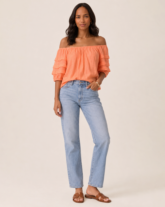 Close-up of a woman wearing a cantaloupe tier sleeve top and light wash denim jeans on a beige background