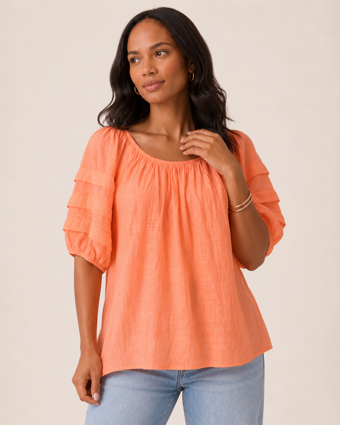 Close-up of a woman wearing a cantaloupe tier sleeve top and light wash denim jeans on a beige background