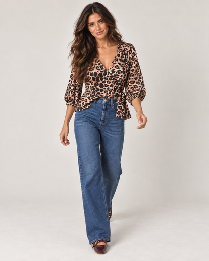 Full-body front view of a woman wearing a leopard print V-neck blouse with long sleeves, brown pants, and dark ankle boots on a beige background.