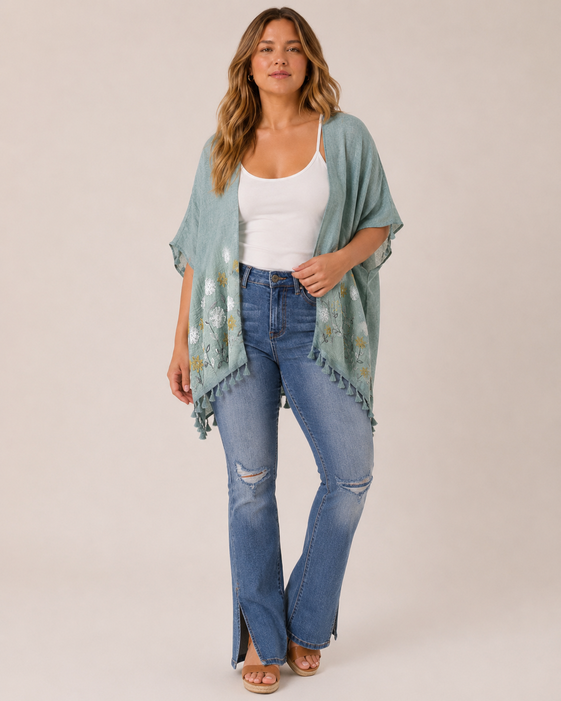 Full-body view of a woman wearing a sage embroidered kimono over a white tank top, distressed blue jeans, and tan sandals on a beige background