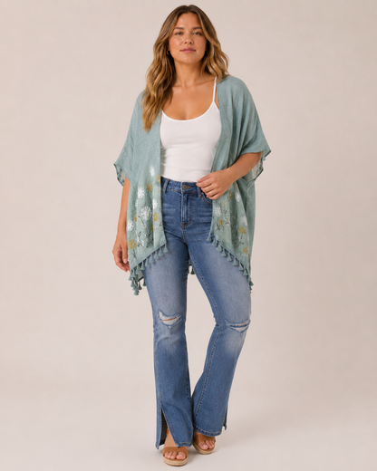 Full-body view of a woman wearing a sage embroidered kimono over a white tank top, distressed blue jeans, and tan sandals on a beige background