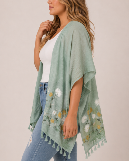 Side view of a woman wearing a sage embroidered kimono over a white tank top and distressed blue jeans on a beige background