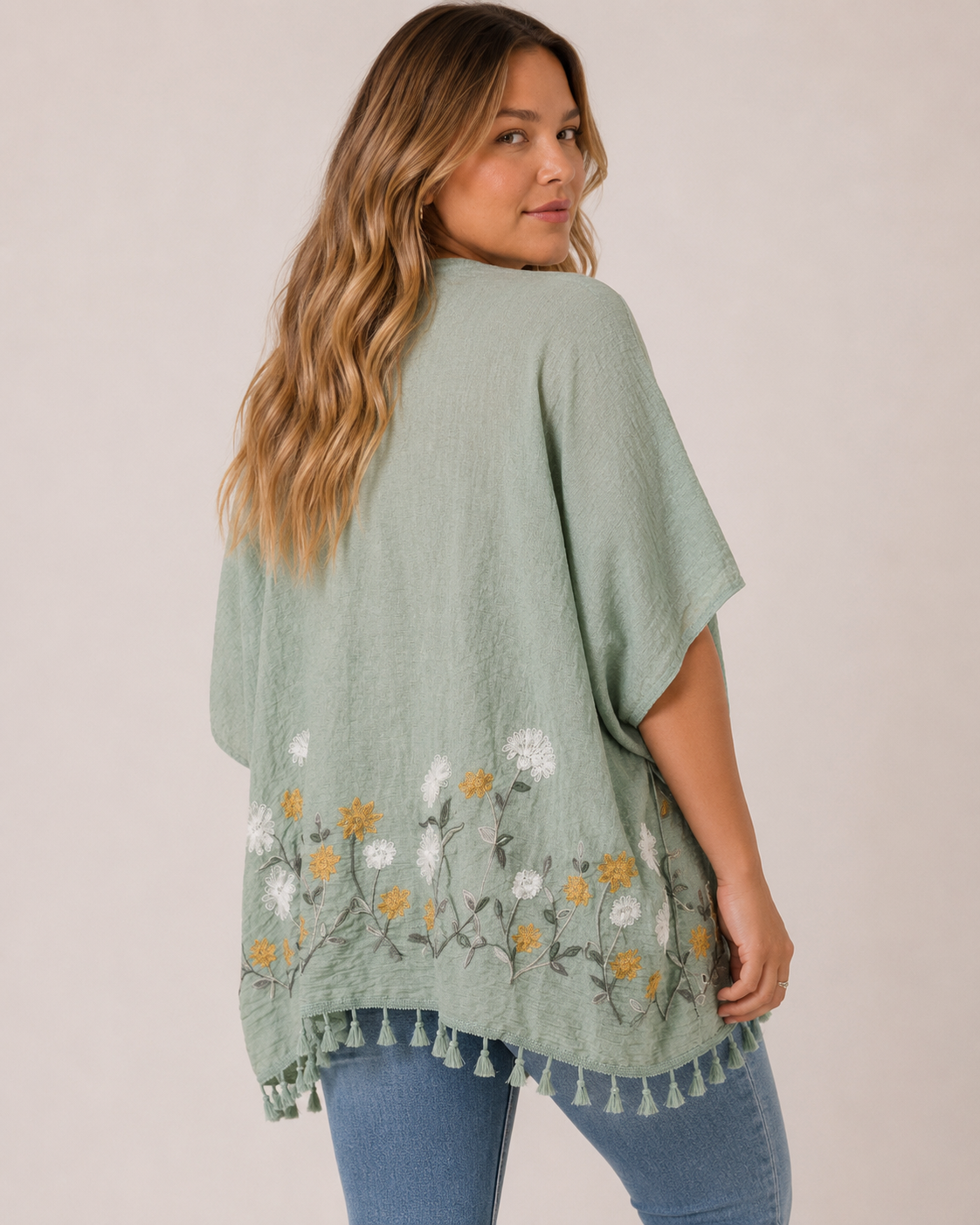 Back view of a woman wearing a sage embroidered kimono over a white tank top and distressed blue jeans on a beige background